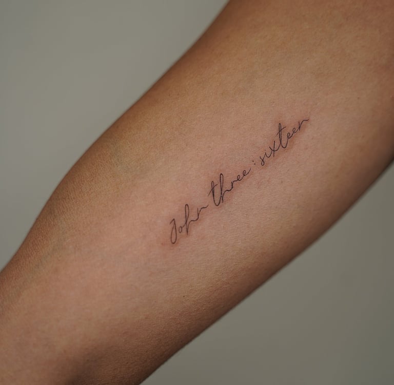 Tatuaje fineline en Madrid de frase John three sixteen en cursiva fina diagonal en el antebrazo