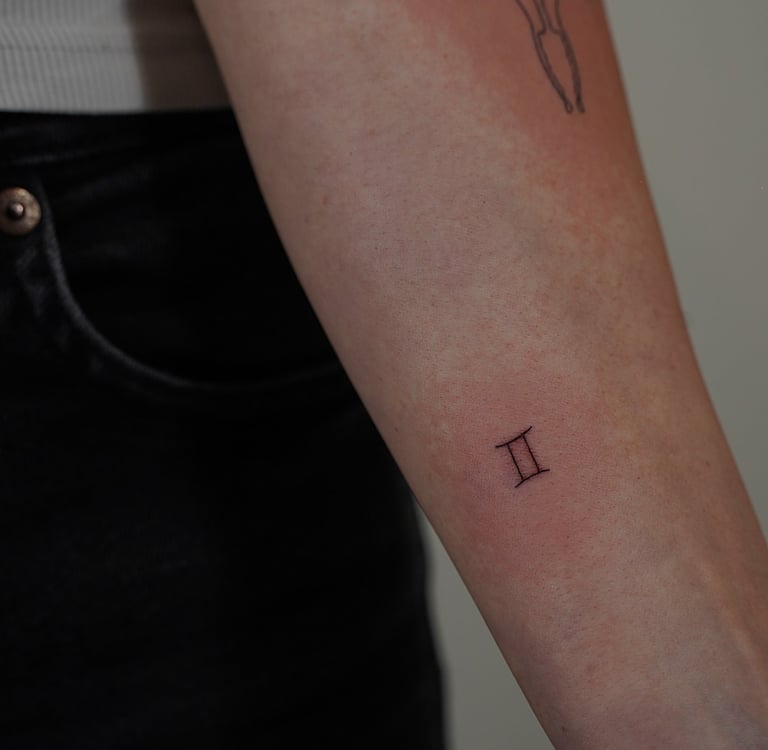 Tatuaje fineline en Madrid de símbolo del zodiaco Géminis en trazo fino en el antebrazo