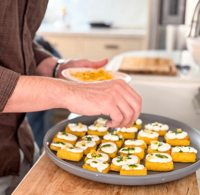 A chef garnishes a tray of crispy polenta squares topped with creamy goat cheese and fresh herbs.