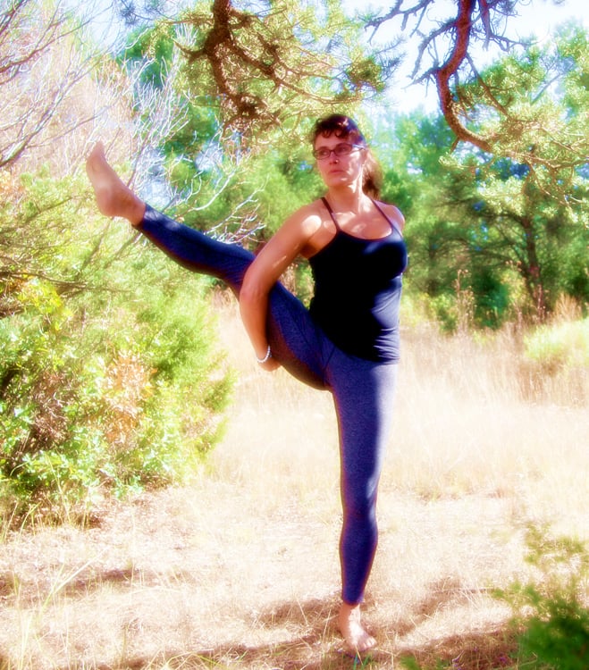 A woman practices a standing balance yoga pose in a sunny forest clearing for outdoor wellness.