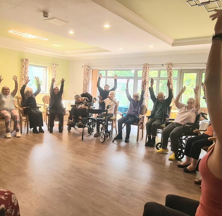 Group of elderly residents taking part in a seated exercise and chair yoga class, led by Lucy