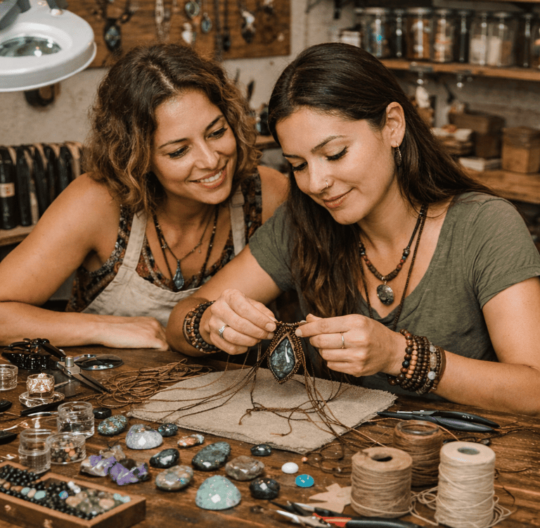 Dos artesanas elaborando joyería de macramé hecha a mano con piedras preciosas en una mesa de taller