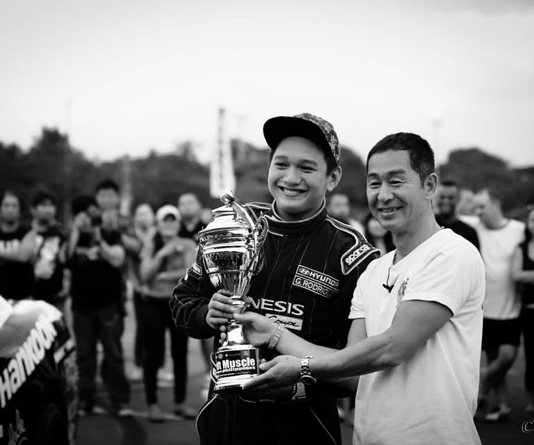 Keiichi Tsuchiya handing a trophy to a man in front of a crowd 