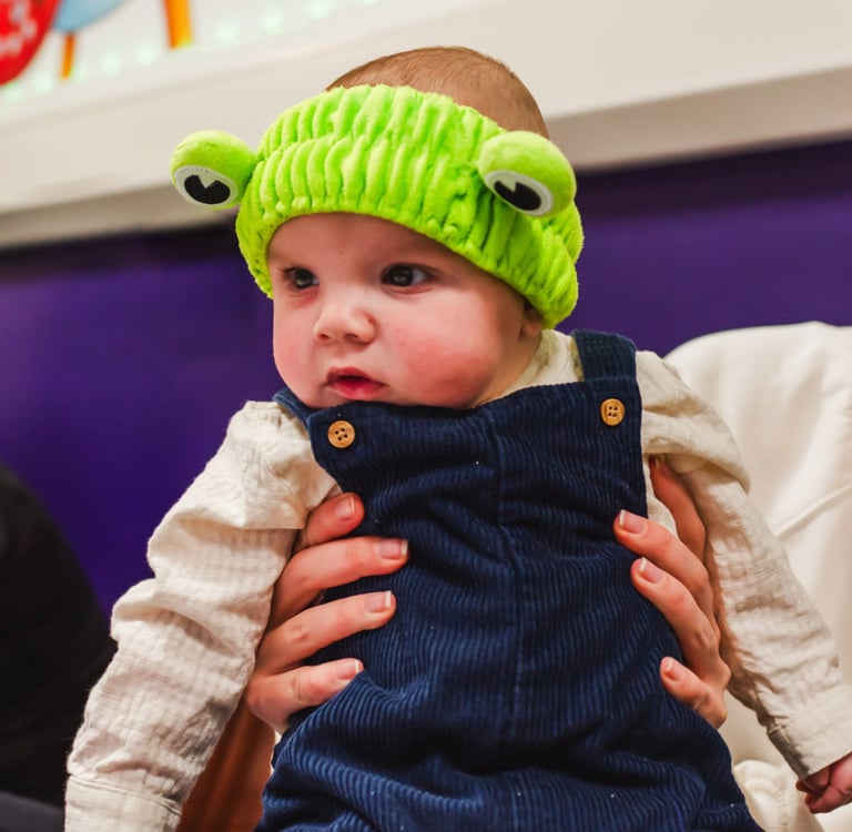 a baby wearing a a frog headband enjoying moo music class with her mum