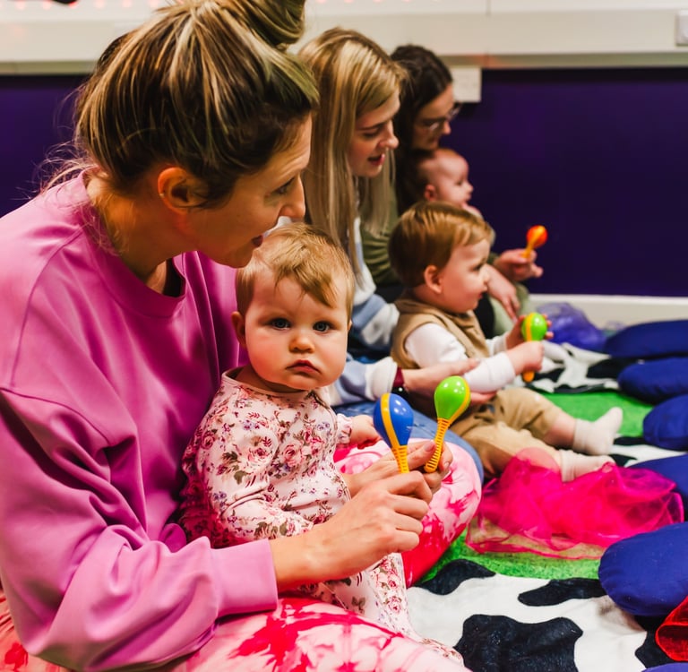 Young girl sat on mums lap playing with shaker instruments. Other children with their mums too.