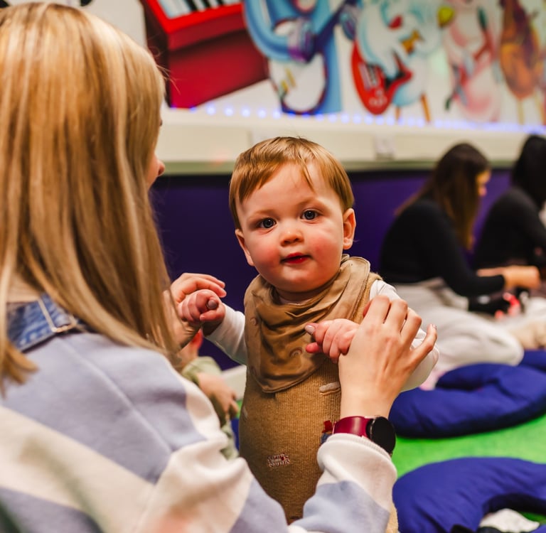 A young boy staring into the camera, holding his mums hands to stand up in moo class.