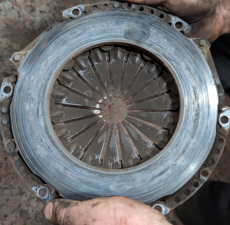 A mechanic holds a worn automotive clutch pressure plate showing signs of friction surface heat damage.