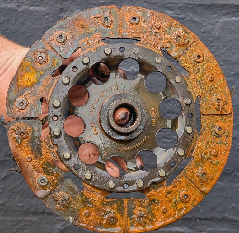 Hand holding a heavily rusted and worn automotive clutch disc against a dark wall background.