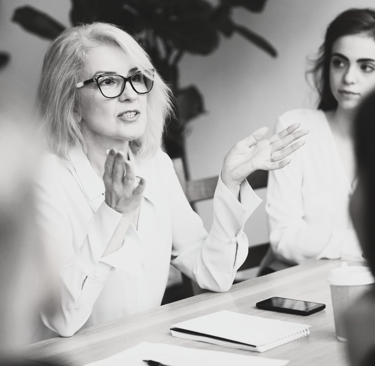 a woman in a white shirt and glasses sitting at a meeting table gesturing with her hands