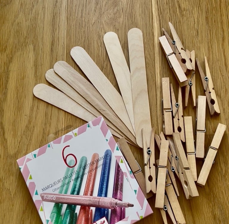 A box of Djeco glitter markers next to wooden craft sticks and clothespins on a table.