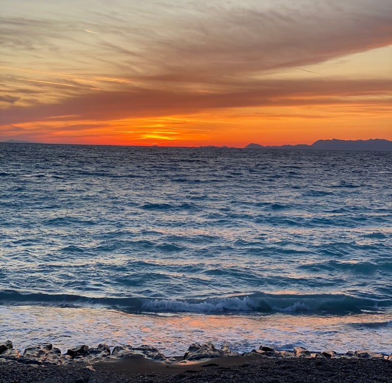 Vibrant orange sunset over blue ocean waves on a rocky beach with distant mountain silhouettes.