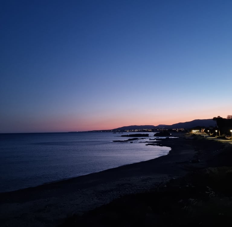A scenic dusk view of a dark sandy beach coastline with distant city lights and a purple sunset horizon.