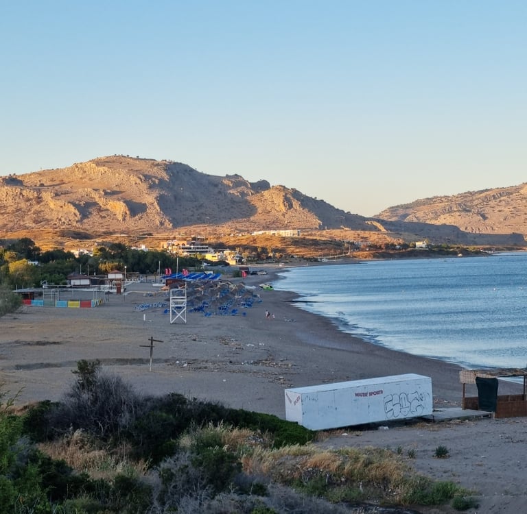 Panoramic view of a sandy Mediterranean beach with sunbeds, blue sea, and rocky mountains at sunset.
