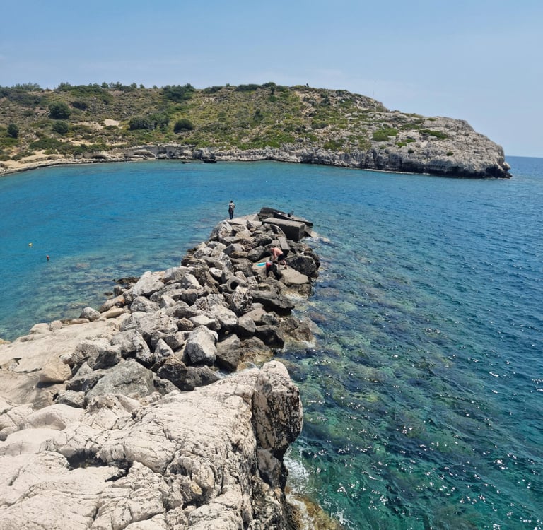 People relax and explore on a rocky breakwater in a beautiful, clear blue Mediterranean cove, perfect for snorkeling and
