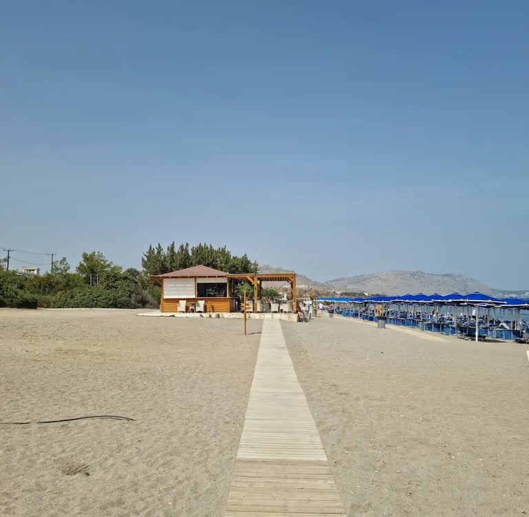 A wooden boardwalk leads to a beach bar with blue umbrellas on a sunny sandy shore.