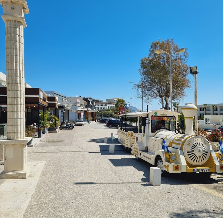 A white and gold tourist fun train parked on a sunny street in a Greek resort town.