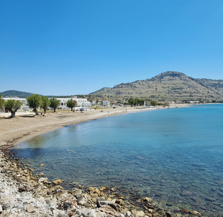 Panoramic view of a sandy Greek beach with turquoise sea, white buildings, and coastal mountains under a clear blue sky.
