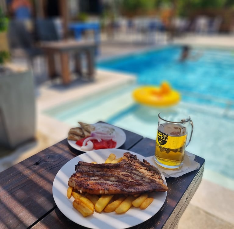 Grilled pork steak and french fries served with cold beer by a sunny swimming pool.