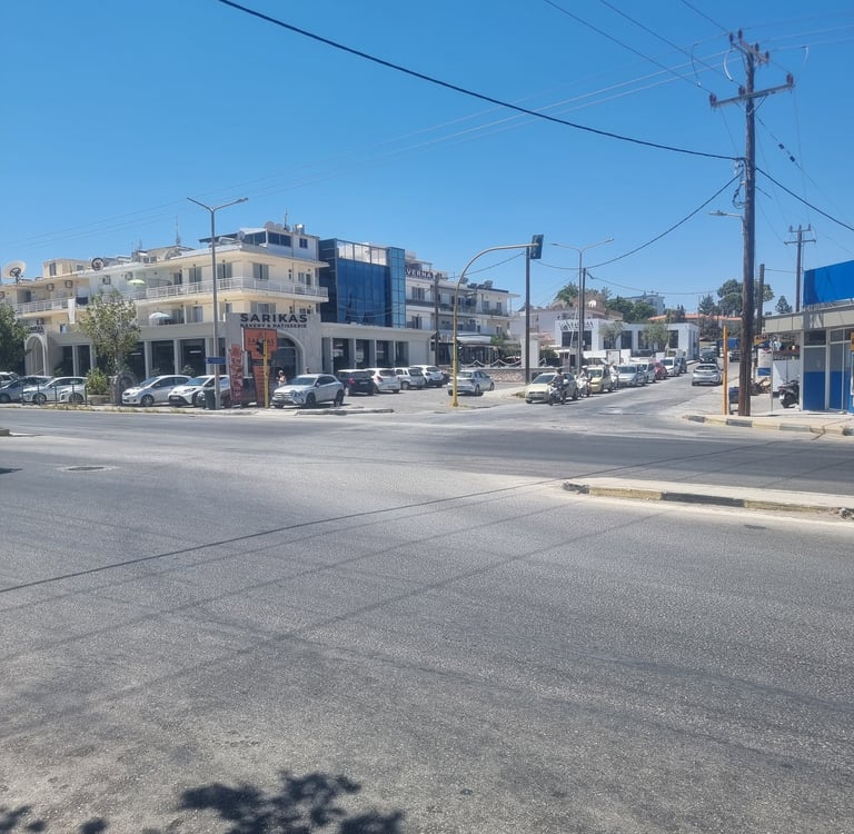 Sunny street intersection in Faliraki Rhodes with parked cars near the Sarikas building.
