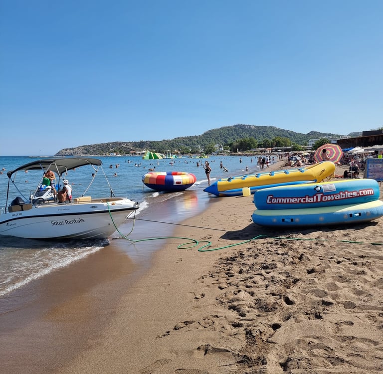 A motorboat and inflatable water sports towables sit on a crowded sandy beach under a clear blue sky.