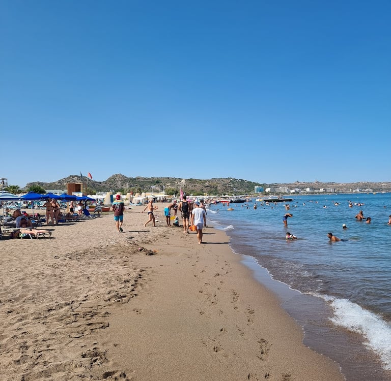 Crowded sandy beach in Rhodes, Greece, with tourists swimming in the blue Mediterranean Sea under a clear sky.