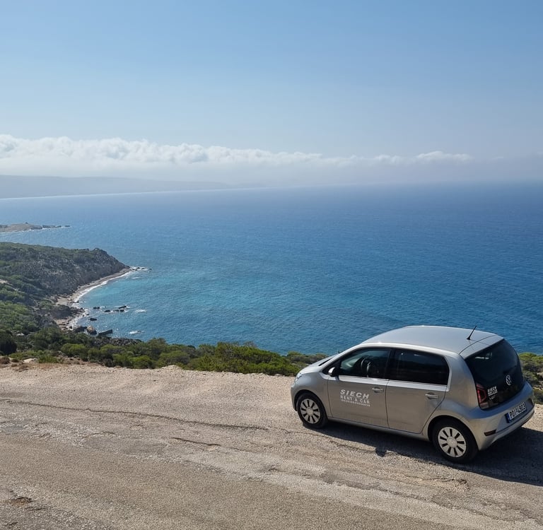 A silver compact rental car parked on a scenic coastal overlook with a view of the blue Mediterranean sea.