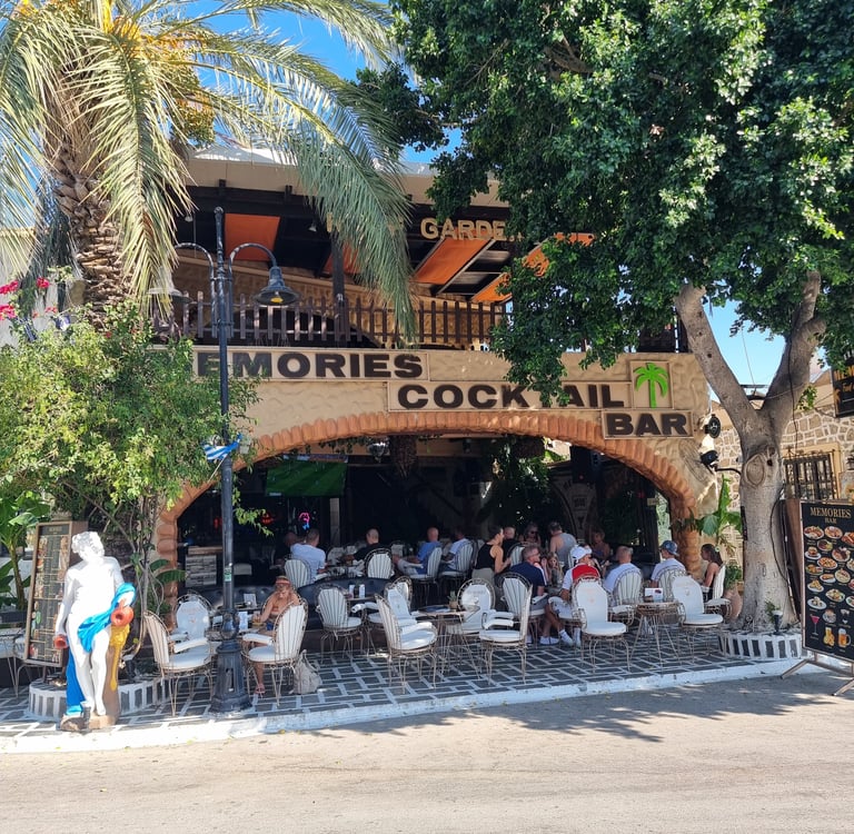 Patrons enjoy drinks at the outdoor Memories Cocktail Bar terrace under palm trees in Greece.