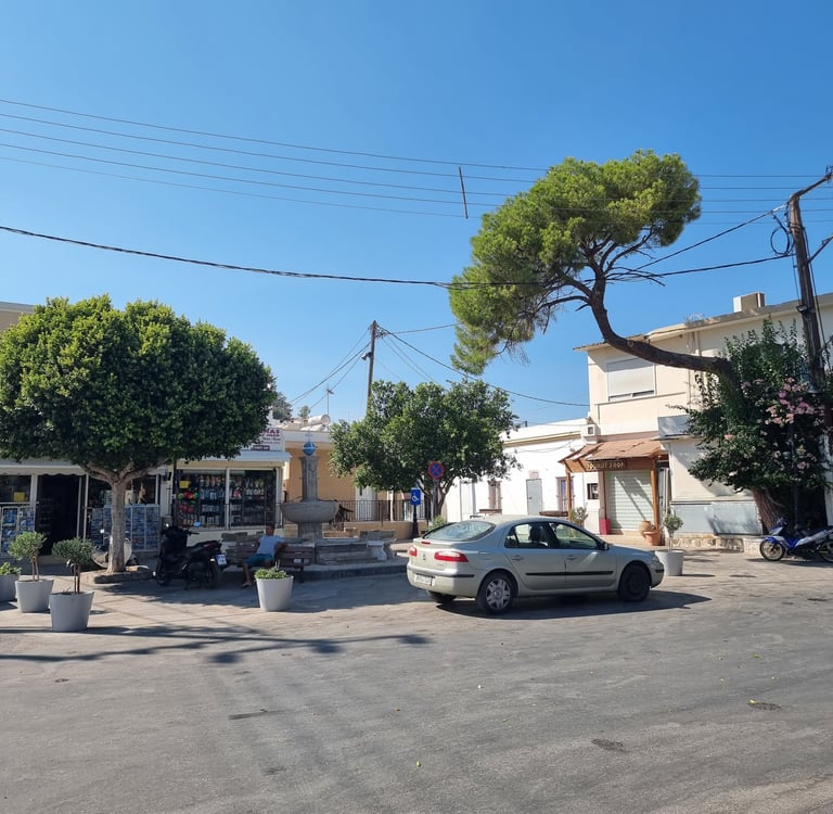 Sunny town square in Greece with a silver car parked near green trees and white buildings.
