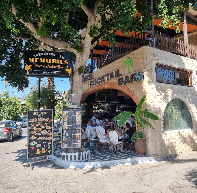 Memories Food and Cocktail Bar featuring an outdoor stone patio with patrons and menu boards under a large tree.