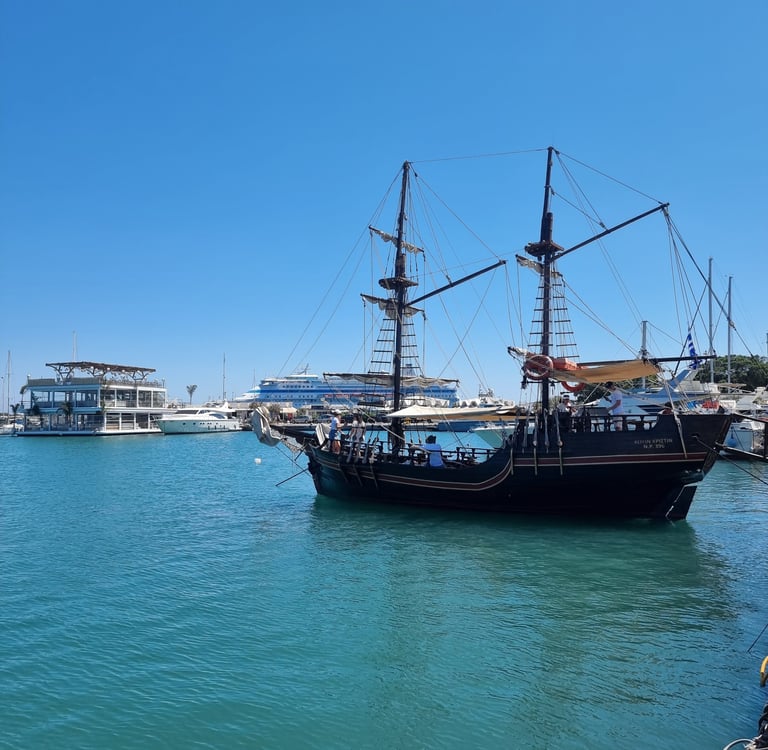 A vintage-style wooden pirate ship tour boat sailing in the blue waters of a Mediterranean harbor.