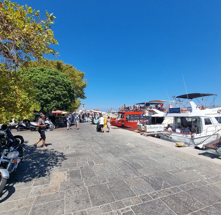 Crowded Mandraki Harbor in Rhodes Greece with tourist boats moored along the stone promenade under a blue sky.