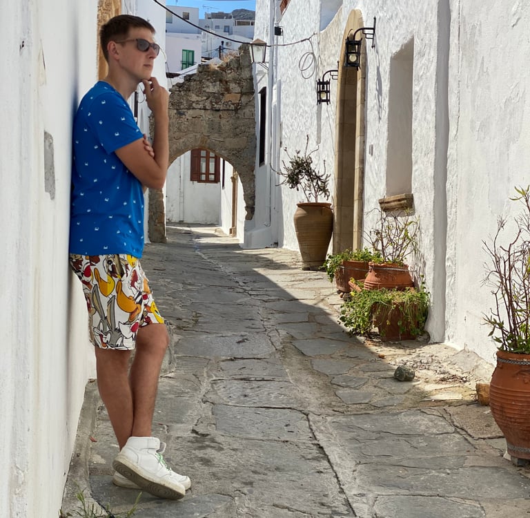 A man in a blue shirt leaning against a white wall in a narrow cobblestone street in Greece.