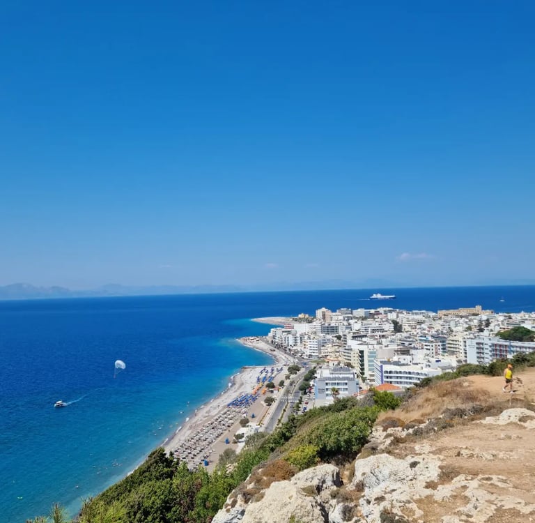 Aerial view of Rhodes City beach with white buildings, blue Aegean Sea, and parasailing on a sunny day.