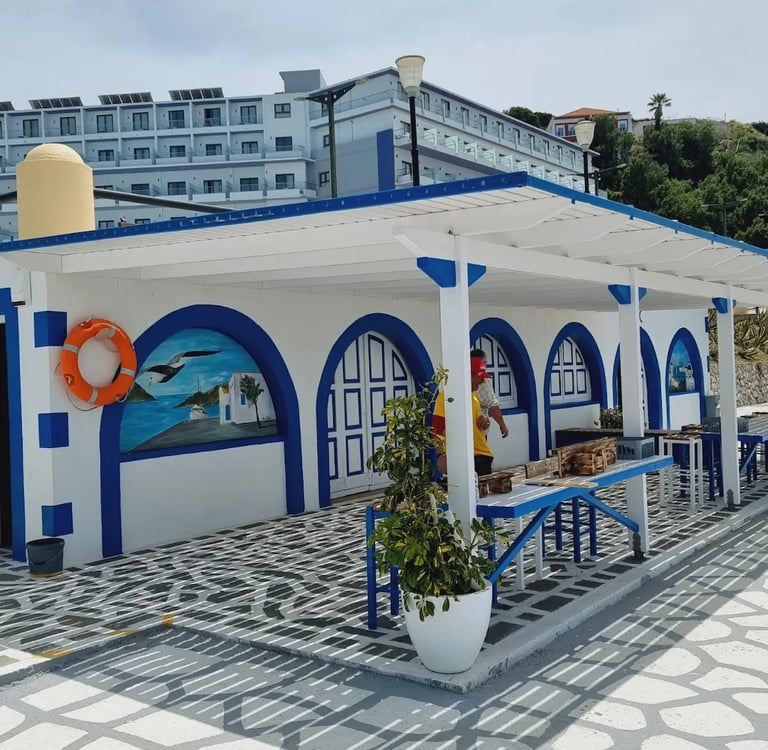 White and blue Greek style seaside building with arched murals and an outdoor patio.