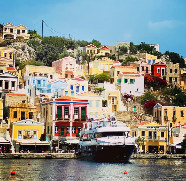 Colorful neoclassical houses on a hillside overlooking a ferry at Symi Island harbor in Greece.