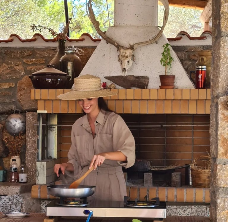 Smiling woman in a straw hat cooking outdoors on a gas stove in a rustic stone kitchen.