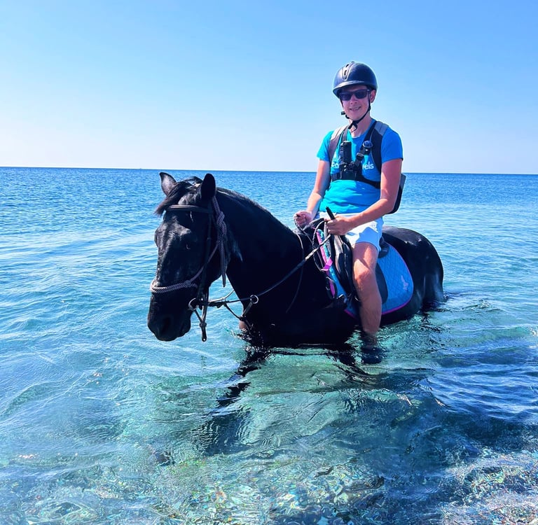 looneytravels riding a black horse through clear blue ocean water during a summer beach horse trek.