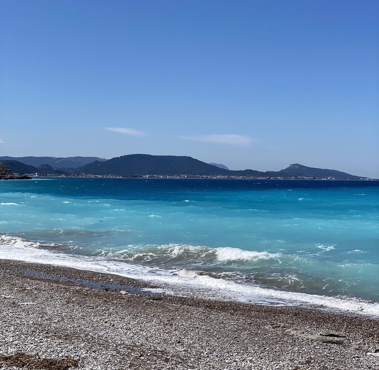 Pebble beach with turquoise Mediterranean waves and mountains under a clear blue sky.