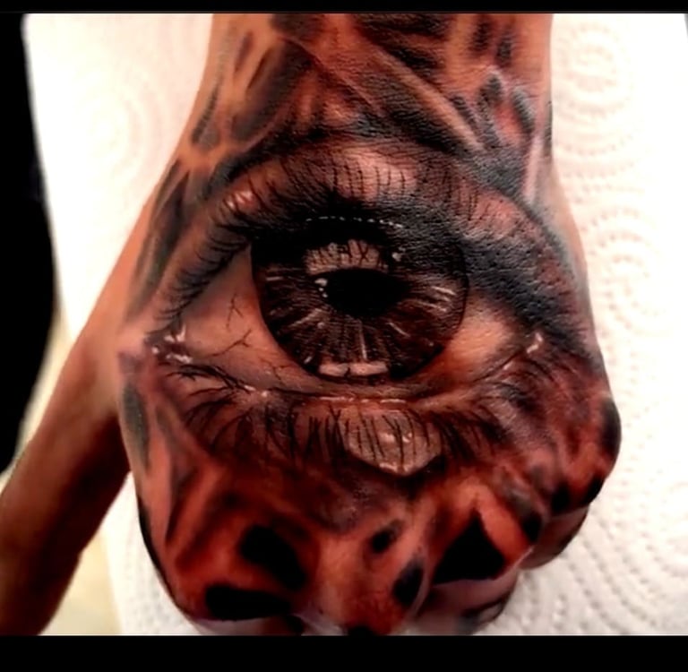 Realistic black and grey eye tattoo on a hand with intricate pupil details and surrounding dark patterns.