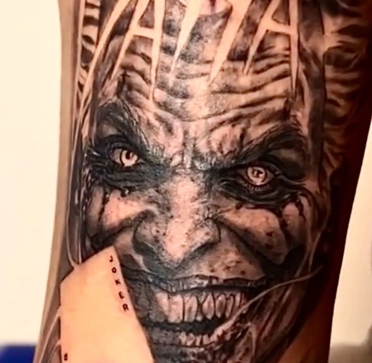 Detailed black and grey Joker tattoo featuring a realistic villainous grin, HA HA text, and a joker playing card.