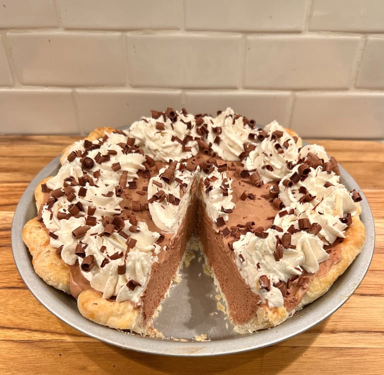 A homemade chocolate silk pie with whipped cream and chocolate shavings on a wooden counter.