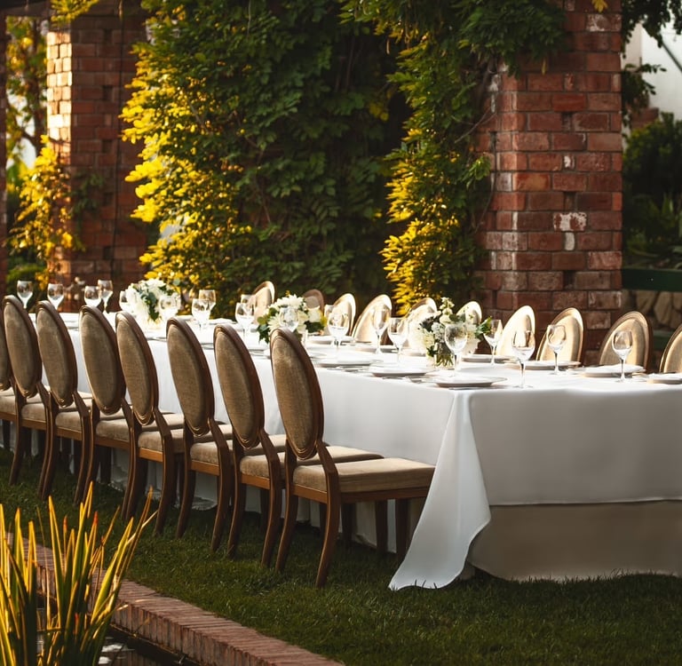 Elegant outdoor banquet table set for a wedding dinner in a lush garden with brick pillars.