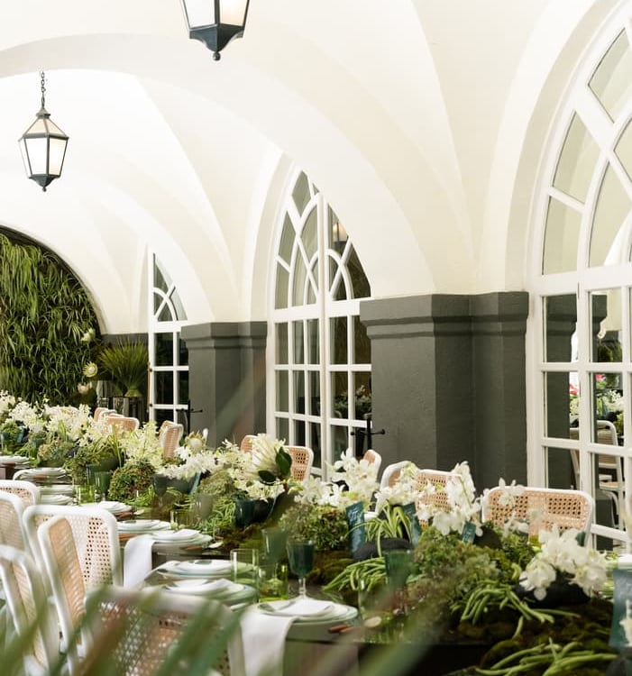 Elegant outdoor dining table setting with green moss runners and white floral centerpieces under arched ceilings.