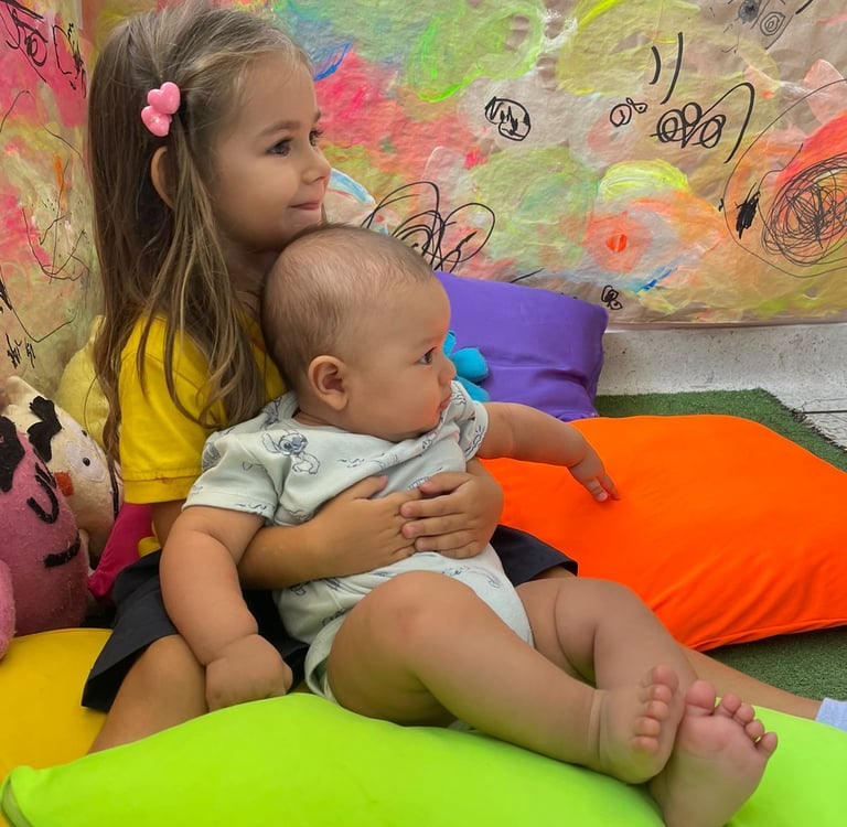 Niña abrazando bebe sentados entre cojines de colores vibrantes en Ukelele Jardín Infantil