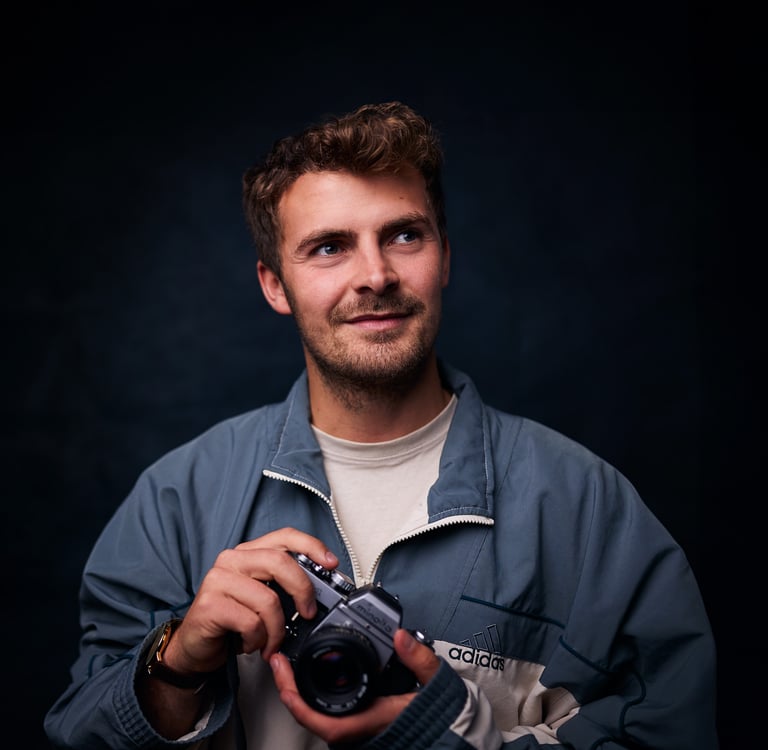 A smiling man holding a vintage Minolta film camera while wearing an Adidas jacket.
