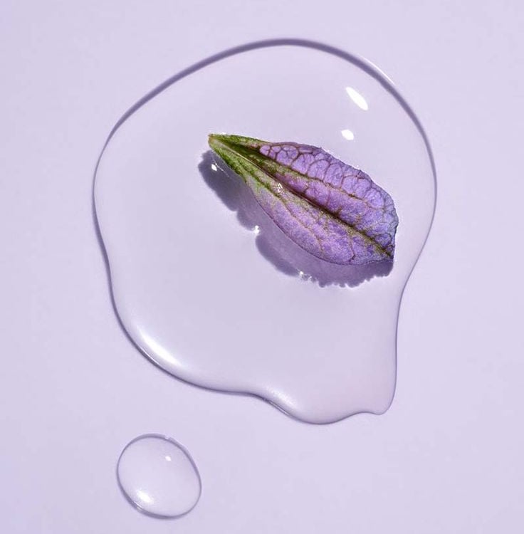 A close-up of a purple flower petal resting on a clear liquid, symbolizing the gentle and hydrating