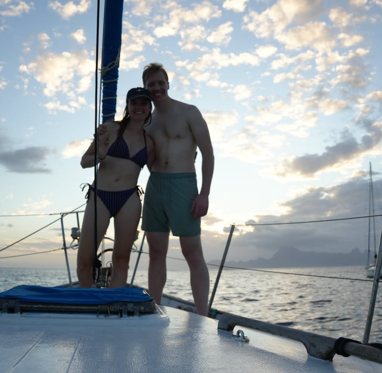 Alex and Niccy smiling at the bow of their sailboat under sunny skies