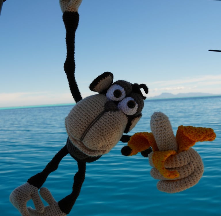 A crochet chimpanzee hanging from a boat rail holding a yarn banana over blue tropical ocean water.