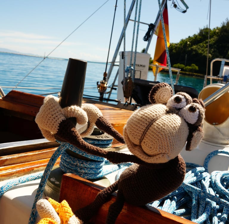 A cute crochet monkey toy holding a winch and banana on a sailboat deck under a clear blue sky.