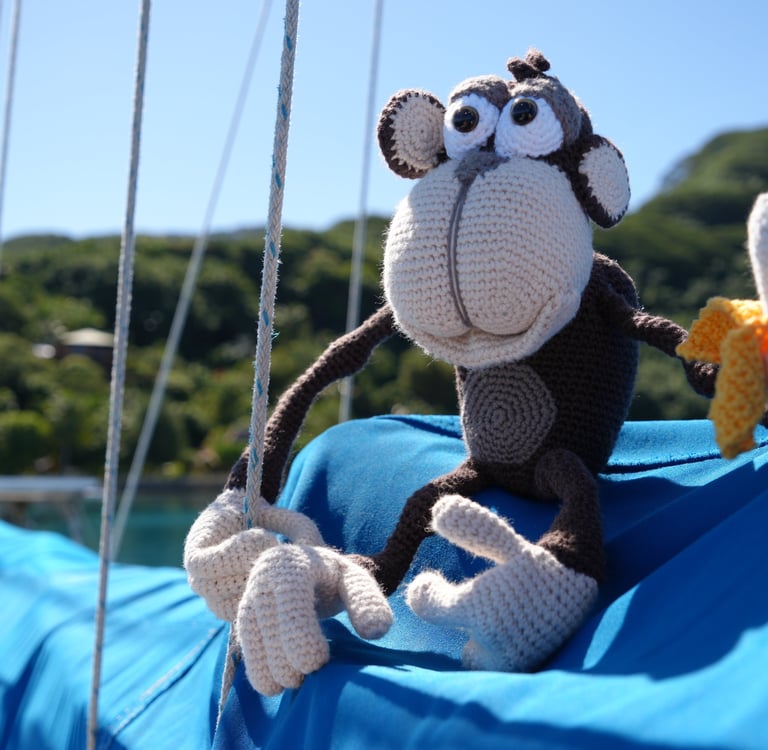A handmade crochet monkey plush toy holding a banana on a blue boat sail with a tropical island background.